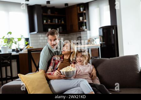 Une famille de trois est confortablement nichée sur un canapé, leurs visages reflétant l'excitation et l'attention comme ils partagent un bol de pop-corn pendant un susp Banque D'Images