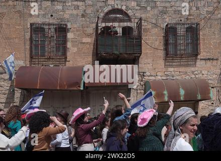 Une palestinienne regarde avec ses enfants depuis le balcon de leurs colons juifs en costumes danser alors qu’ils marchent dans la rue Al-Shuhada, qui est largement fermée aux Palestiniens, pendant la parade annuelle de Pourim alors que les forces de sécurité israéliennes sécurisent les célébrations du 24 mars 2024 à Hébron, en Israël. Banque D'Images