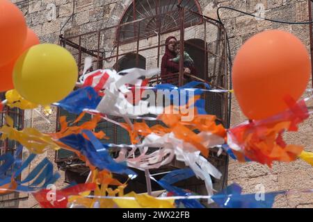 Une palestinienne regarde avec ses enfants depuis le balcon de leurs colons juifs en costumes danser alors qu’ils marchent dans la rue Al-Shuhada, qui est largement fermée aux Palestiniens, pendant la parade annuelle de Pourim alors que les forces de sécurité israéliennes sécurisent les célébrations du 24 mars 2024 à Hébron, en Israël. Banque D'Images