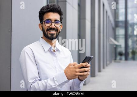 Portrait d'un jeune Indien souriant et heureux dans une chemise blanche et un short debout près d'un centre de bureau, tenant un téléphone et souriant à la caméra. Banque D'Images