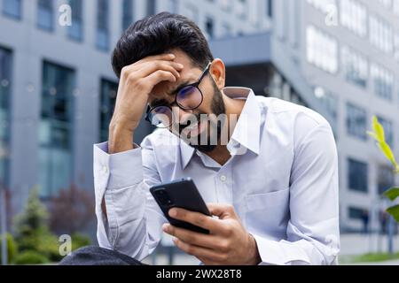 Photo en gros plan d'un jeune homme indien choqué et bouleversé assis à l'extérieur tenant la tête avec la main et regardant l'écran du téléphone après avoir reçu de mauvaises nouvelles. Banque D'Images