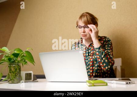 Une jeune femme blonde portant des lunettes et utilisant un ordinateur portable à la maison, se concentrant sur l'écran, représentant le travail à distance ou l'étude, adapté pour en ligne Banque D'Images