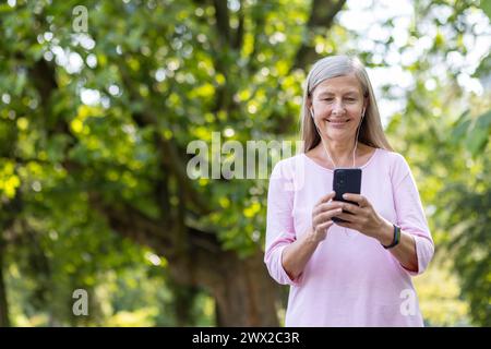 Femme âgée souriante en t-shirt rose debout à l'extérieur dans le parc portant un casque et utilisant un téléphone portable, discutant, tapant un message. Banque D'Images
