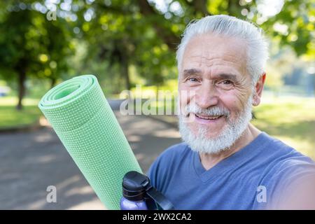 Photo en gros plan d'un homme aux cheveux gris plus âgé souriant debout dans un parc, tenant une natte et une bouteille d'eau, souriant et parlant à la caméra du téléphone. Banque D'Images