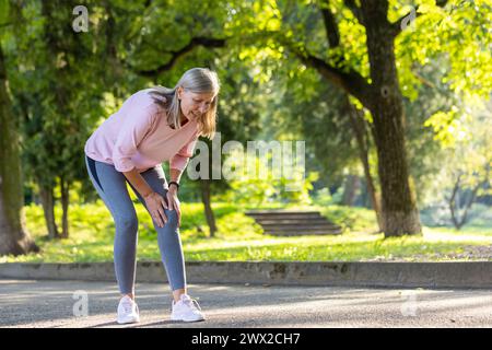 Femme âgée aux cheveux gris marche et fait du sport dans le parc, se tient penchée dans la douleur, tient les mains sur le genou. Banque D'Images