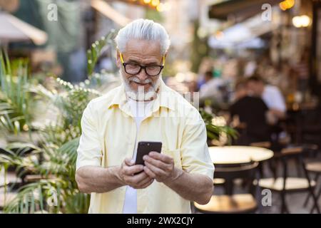 Un homme âgé heureux barbu aux cheveux gris dans une chemise jaune et des lunettes marche dans la ville et utilise un téléphone portable avec le sourire. Banque D'Images