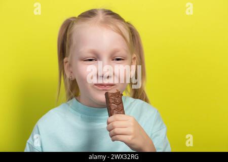 Une belle fille joyeuse aux cheveux blancs de sept ans mange une barre de chocolat sur fond jaune. Les enfants sont des amoureux de la malbouffe sucrée, horizonto Banque D'Images