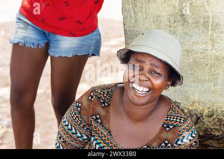 femme villageoise africaine célibataire portant un chapeau, assise dehors devant la maison dans un village, Banque D'Images