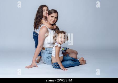 Belle mère latina avec de longs cheveux bruns en vêtements blancs et bleus, assise avec ses deux jolies petites filles avec des boucles vénézuéliennes, photo studio Banque D'Images