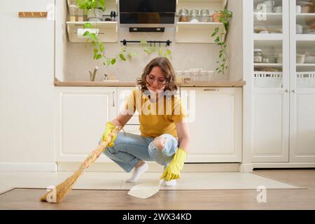 Une femme balaie le sol avec un balai et une pelle à poussière dans sa cuisine. Mains féminines en gants jaunes lors du nettoyage de la cuisine Banque D'Images