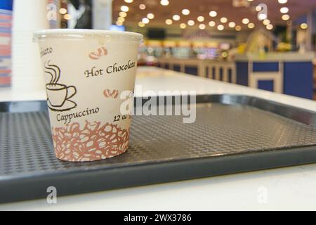 Gobelet en papier avec des inscriptions de café et de chocolat chaud, placé sur un plateau noir sur un comptoir, avec un fond flou d'un café éclairé. Banque D'Images