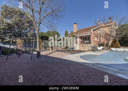 Terrain détaché de type chalet avec sol en gravier, quelques cyprès à l'intérieur, piscine couverte de toile bleue et balançoires pour enfants en métal gris Banque D'Images