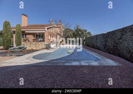 Terrain de chalet détaché avec des planchers de gravier, quelques cyprès à l'intérieur, une piscine couverte de toile bleue et une clôture de périmètre avec des haies taillées Banque D'Images