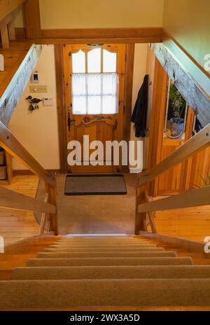 Escalier raide avec des marches recouvertes de tapis bronzé menant au rez-de-chaussée et à la porte d'entrée principale à l'intérieur de la maison de chalet en rondins de style Canadiana, Québec, Canada Banque D'Images