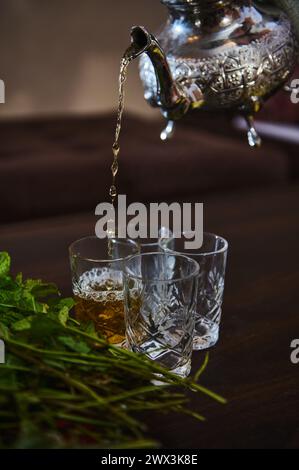 Verser un délicieux thé à la menthe marocain d'une théière en argent dans des verres à boire. Bouquet de menthe fraîche au premier plan Banque D'Images