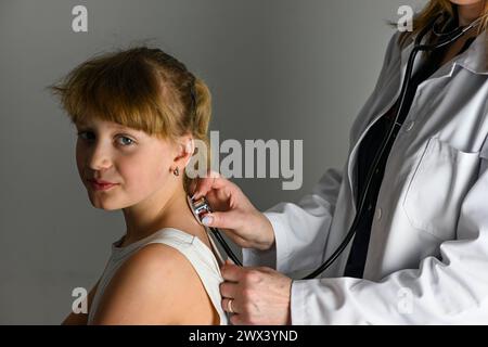 un médecin en manteau blanc avec un stéthoscope écoute la respiration et les poumons de la petite fille. Banque D'Images