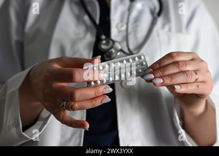 un médecin dans un manteau blanc avec un stéthoscope tient des pilules contraceptives dans sa main, fond flou. Banque D'Images