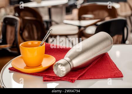 Une bouteille en métal isolée sous vide pour l'eau ou le café et une tasse de café sur une table dans un café. Banque D'Images