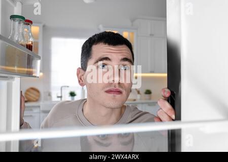 Homme bouleversé près d'un réfrigérateur vide dans la cuisine, vue de l'intérieur Banque D'Images