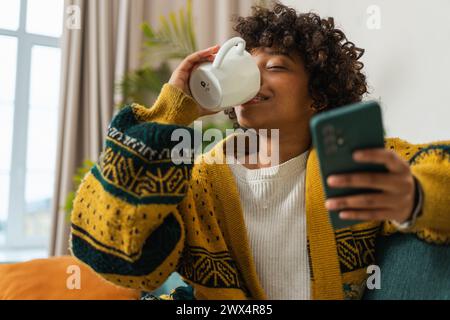 Bonjour. Fille africaine buvant du café tenant un smartphone assis sur un canapé à la maison femme avec téléphone portable surfant sur Internet en utilisant des applications de médias sociaux. Acheter en ligne Internet nouvelles dépendance au téléphone portable Banque D'Images