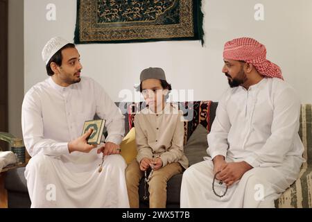 Hommes musulmans modernes et garçon assis sur le canapé dans le salon discutant suwar du Coran lors d'une réunion de famille sur la fête islamique Banque D'Images