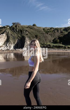 jolie femme blonde souriante en vêtements de sport se tient sur une plage avec des falaises et un ciel bleu en arrière-plan Banque D'Images