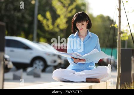 Concentré femme d'affaires entrepreneur lisant des documents et de la documentation, préparant CV CV assis sans chaussures avec dossier sur le banc en milieu urbain Banque D'Images