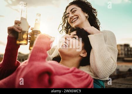 Deux femmes riant avec des bières - célébration du coucher du soleil, chaleur de l'heure d'or - plaisir en plein air insouciant, bonheur franc. Banque D'Images