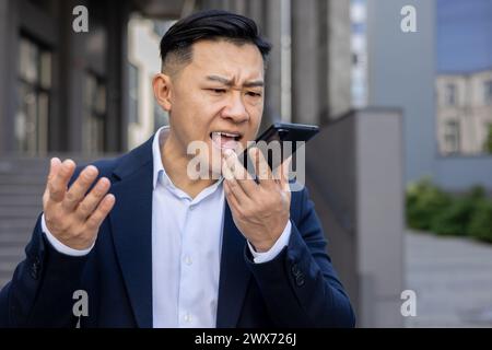 Photo en gros plan de jeune homme d'affaires asiatique en colère en costume debout à l'extérieur et parlant émotionnellement au téléphone à travers le haut-parleur, message vocal insatisfait d'enregistrement. Banque D'Images