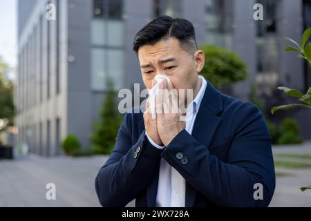 Photo en gros plan d'un jeune homme d'affaires asiatique malade assis sur un banc près d'un immeuble de bureaux et essuyant son nez, avec une serviette d'un nez qui coule. Banque D'Images