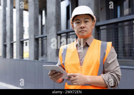 Portrait d'un jeune ingénieur de construction asiatique debout près d'un immeuble de bureaux portant un casque de sécurité et un gilet réfléchissant, tenant un ordinateur portable et regardant sérieusement l'appareil photo. Banque D'Images