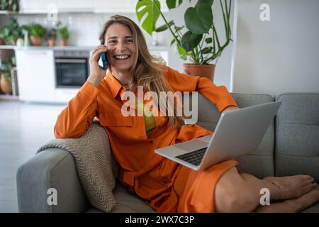 Heureuse femme détendue souriante parlant sur smartphone assis avec ordinateur portable sur canapé avoir un dialogue agréable Banque D'Images