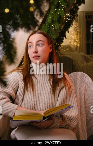 Bonjour l'hiver. femme moderne souriante en pull blanc assis sur la chaise et lisant un livre dans la chambre décorée pour célébrer la nouvelle année et noël Banque D'Images