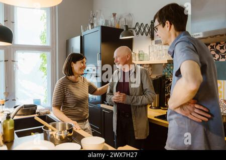 Femme souriante et homme parlant avec le beau-père senior tout en se tenant dans la cuisine à la maison Banque D'Images