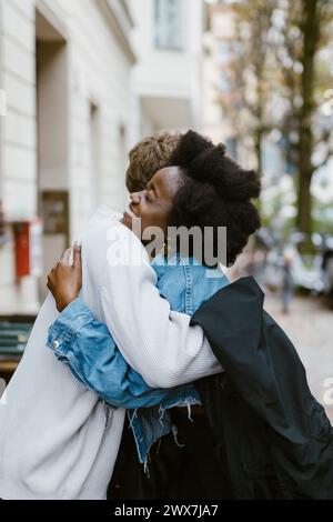 Vue latérale de la femme heureuse embrassant petit ami avec bras autour tout en se tenant sur le trottoir dans la ville Banque D'Images