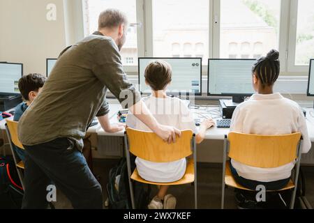 Enseignant masculin aidant les élèves à surfer sur Internet à travers des ordinateurs en classe à l'école Banque D'Images