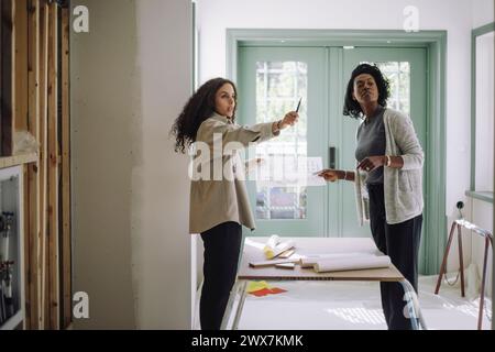 Vue latérale du professionnel de la conception féminine expliquant le plan au client à l'appartement en construction Banque D'Images