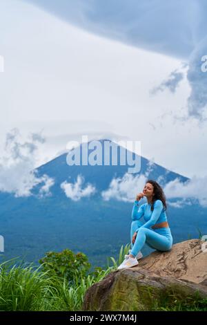 Une jeune femme est assise sur un grand rocher dans un point de vue et profite de la vue sur le volcan sacré du mont Agung caché par les nuages un jour de pluie sur l'île Banque D'Images