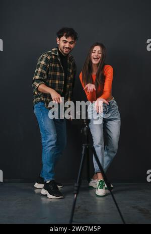 Un jeune couple habillé de façon décontractée, exécutant de la danse contemporaine, montrant des mouvements de danse, utilisant un téléphone portable sur trépied, debout sur fond noir Banque D'Images