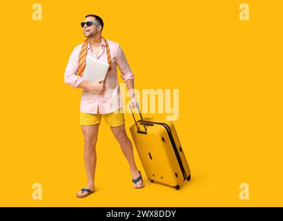 Travailleur de bureau heureux dans des lunettes de soleil avec ordinateur portable et valise sur fond jaune Banque D'Images