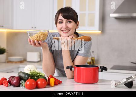 Heureuse jeune femme au foyer avec cuillère et pâtes crues à la table en marbre blanc dans la cuisine. Procédé de cuisson Banque D'Images