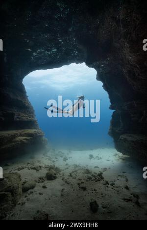 Une jeune femme plongeant librement dans une porte de grotte dans les eaux bleues claires d'Hawaï encadrées par des murs de grotte ombragés. Version de modèle disponible Banque D'Images