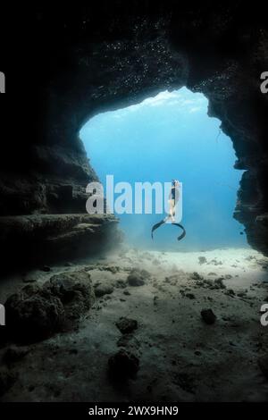 Une jeune femme plongeant librement dans une porte de grotte dans les eaux bleues claires d'Hawaï encadrées par des murs de grotte ombragés. Version de modèle disponible Banque D'Images