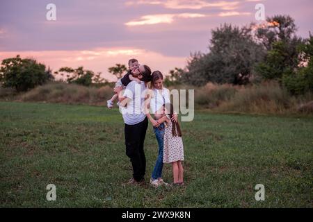 Diversité Happy Family joue sur le terrain au coucher du soleil. Jeune père soulevant le tout-petit haut dans les airs comme la fille aînée et la mère regardent avec charcuterie Banque D'Images