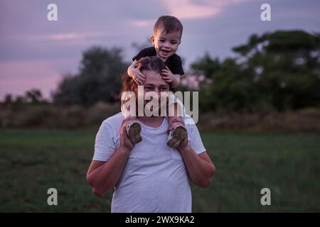 Diversité père avec la barbe portant son fils sur les épaules partage moment tendre dans le champ éclairé crépusculaire, incarnant l'essence de l'amour paternel. Homme et garçon Banque D'Images