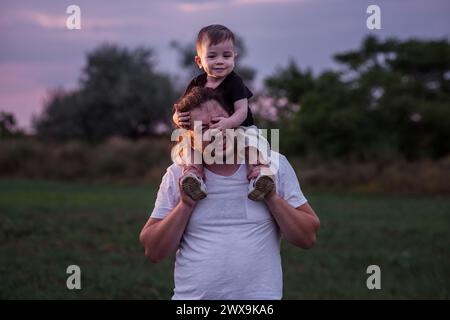 Diversité père avec la barbe portant son fils sur les épaules partage moment tendre dans le champ éclairé crépusculaire, incarnant l'essence de l'amour paternel. Homme et garçon Banque D'Images