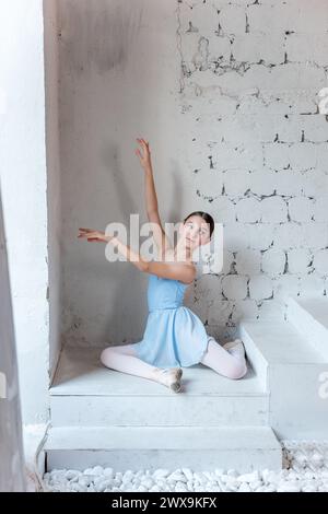 Jeune ballerine poissée en robe bleue crée une pose délicate sur des marches texturées blanches, son élégance contrastant avec la toile de fond rugueuse. Danseurs de ballet po Banque D'Images