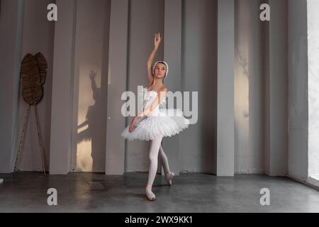 Jeune ballerine apaisée avec diadème à plumes, tutu blanc classique, se tient sur la pointe des pieds à pointe, frappant une pose élégante dans un studio de danse très éclairé, s. Banque D'Images