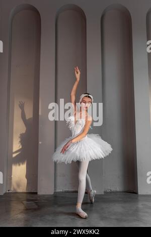 Jeune ballerine apaisée avec diadème à plumes, tutu blanc classique, se tient sur la pointe des pieds à pointe, frappant une pose élégante dans un studio de danse très éclairé, s. Banque D'Images