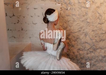 Ballerine contemplative, vêtue de son costume blanc plumeux, est perdue dans la pensée contre un mur texturé, tenant ses chaussures pointe bien usées dans une maman Banque D'Images